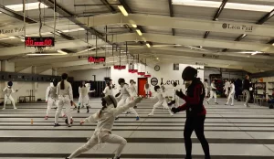 Marion Nemitz giving an individual lesson to a young foilist. There are fencers sparring in the background.