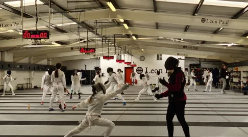 Marion Nemitz giving an individual lesson to a young foilist. There are fencers sparring in the background.