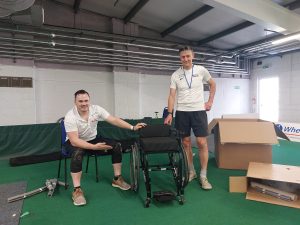 Two BF staff, one a wheelchair user, proudly beside a new RMA fencing wheelchair which is sleek black, an open box is in the image behind and to the right of them indicating the wheelchair has just been set up brand new. 