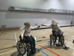 Two fencers sitting in the new RMA Sport Fencing club wheelchairs, their swords crossed and their faces covered by fencing masks