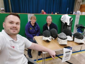 Rick, Alice, and Cheryl sitting at a table with fencing masks and a sign up qr code, all are smiling