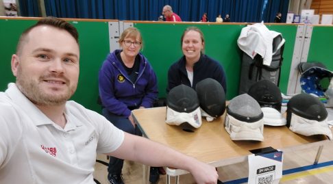 Rick, Alice, and Cheryl sitting at a table with fencing masks and a sign up QR code, all are smiling.