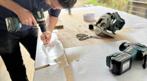 A man building a Sword Seat from a pattern set out on a large piece of wood, there are other tools around him but he is currently using a drill